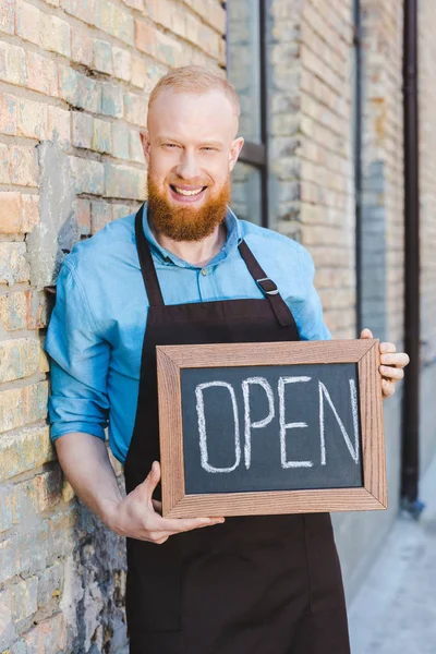 Handsome bearded young barista holding sign open and smiling at camera — Stock Photo