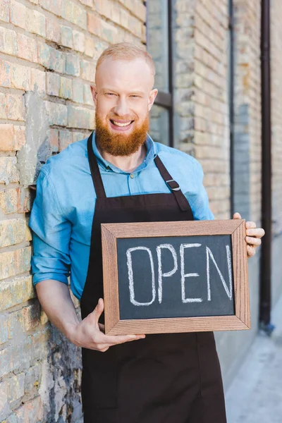 Schöner bärtiger junger Barista hält Schild offen und lächelt in die Kamera — Stockfoto