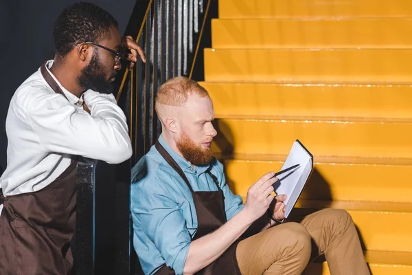 Two multiethnic owners of coffee shop looking at notebook and discussing business on stairs — Stock Photo