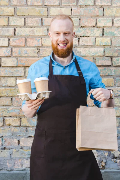 Sonriente joven barista en delantal sosteniendo tazas de papel de café en bolsas de cartón y papeles - foto de stock