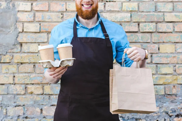 Tiro recortado de barista masculino sonriente en delantal sosteniendo bolsas de papel y tazas desechables con café - foto de stock