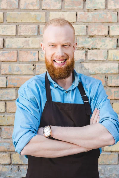 Portrait of young smiling male barista in apron standing in front of brick wall — Stock Photo