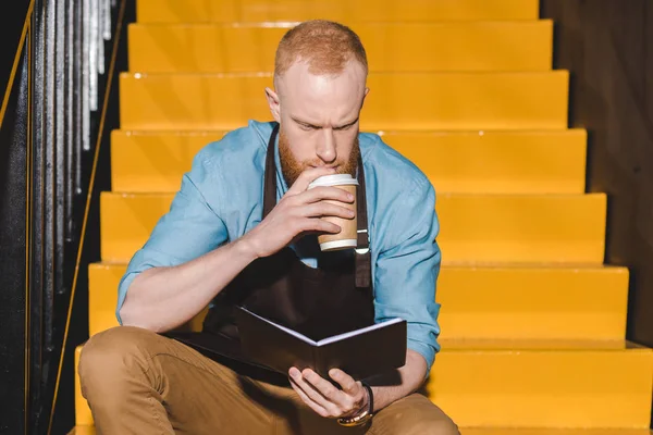 Jovem barista masculino no avental beber café de copo de papel e olhando no livro didático — Fotografia de Stock