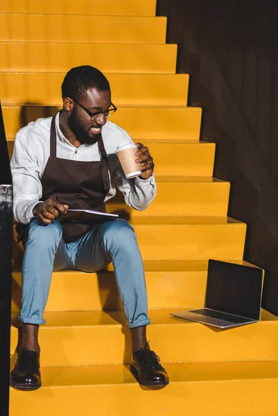 Sorridente afroamericano barista maschile con libro di testo bere caffè e seduto sulle scale con computer portatile — Foto stock