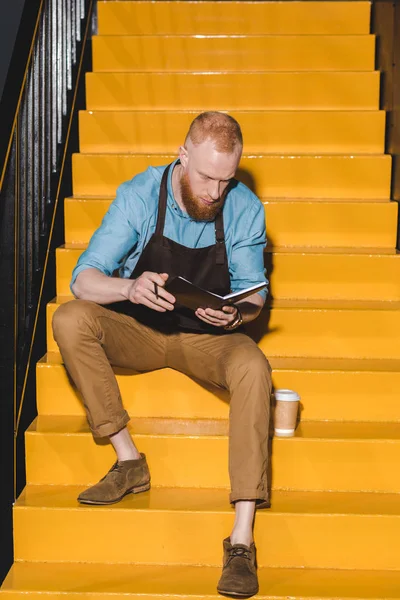 Joven barista en delantal sosteniendo libro de texto y sentado en escaleras con taza de café desechable - foto de stock