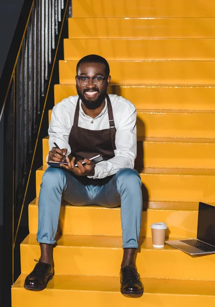 Молодих афро-американських чоловіків barista в фартух холдингу підручник посміхається і сидячи на сходах ноутбук і паперу чашкою ароматної кави — стокове фото