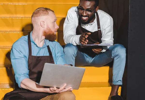 Male barista showing laptop screen to young colleague on stairs — Stock Photo
