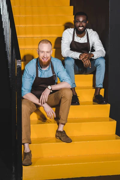 Dois sorridentes proprietários multiétnicos de café em aventais sentados em escadas — Fotografia de Stock