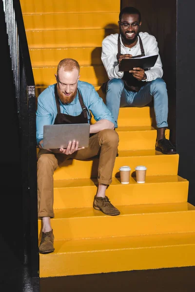 Zwei junge lächelnde Coffeeshop-Besitzer in Schürzen mit Laptop und Lehrbuch sitzen auf der Treppe mit Pappbechern Kaffee — Stockfoto