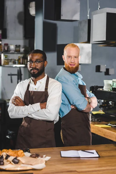 Dos jóvenes baristas multiculturales en delantales de pie con los brazos cruzados en la cafetería - foto de stock