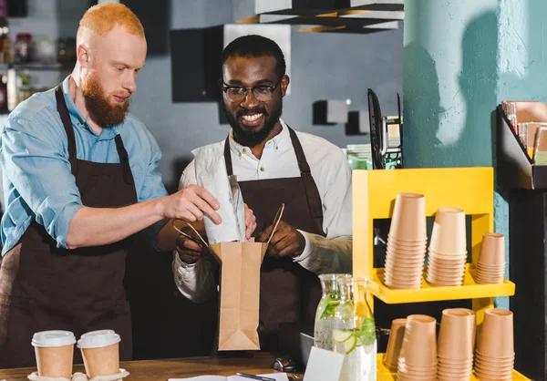 Sonriendo jóvenes propietarios multiétnicos de cafetería poniendo orden en bolsa de papel - foto de stock
