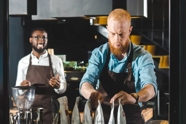 Young male barista taking packages and smiling colleague standing behind in coffee shop — Stock Photo
