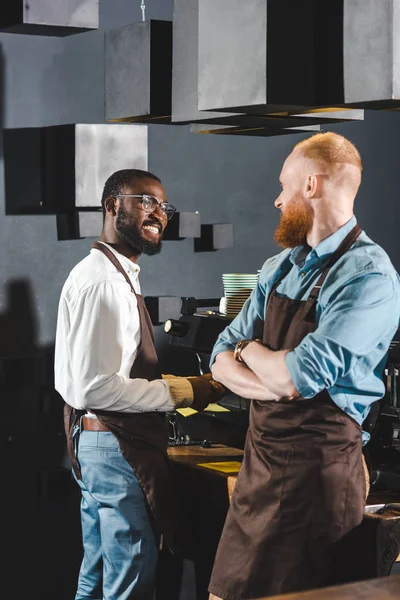 Young smiling multiethnic owners of coffee shop in aprons talking in coffee shop — Stock Photo