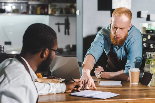 Young serious male barista pointing by finger on textbook to colleague — Stock Photo