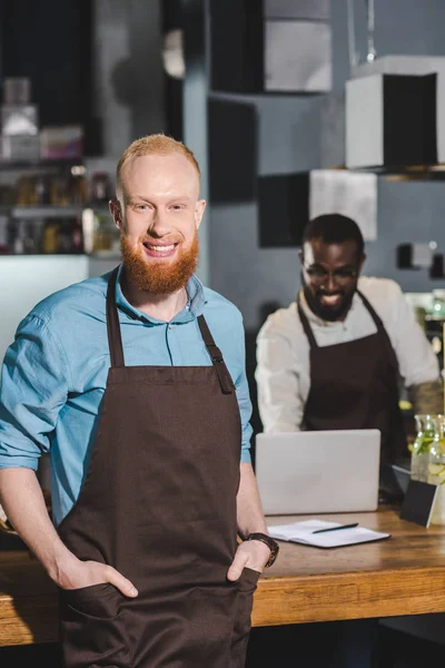 Sorridente jovem barista masculino no avental com colega no café — Fotografia de Stock
