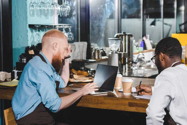 Rear view of young male multiethnic owners of coffee shop working at table with textbook and laptop — Stock Photo