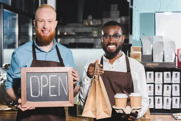 Lächelnde junge multiethnische Coffeeshop-Besitzer mit Kaffeetassen, Papiertüten und Tafel mit aufgeklapptem Schriftzug — Stockfoto