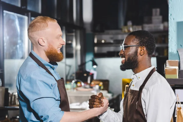 Vue latérale de deux jeunes propriétaires multiethniques de café dans des tabliers serrant la main dans un café — Photo de stock