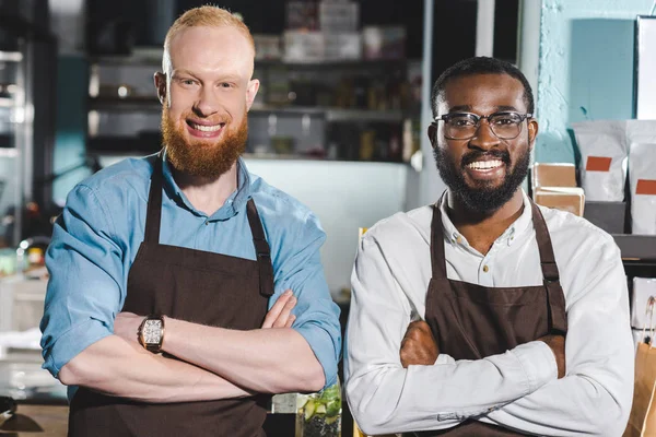 Retrato de dos jóvenes propietarios multiétnicos de cafetería en delantales de pie con los brazos cruzados en la cafetería - foto de stock