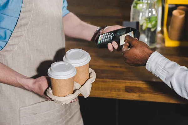 Cropped shot of client paying by credit card and barista with terminal and coffee cups in hands — Stock Photo