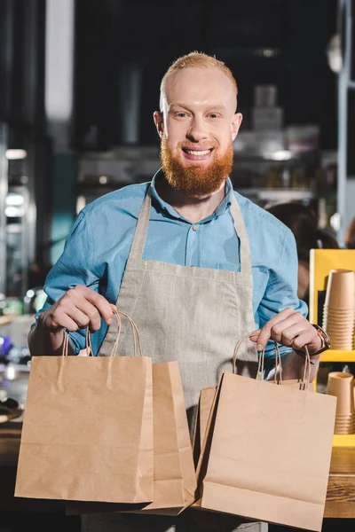 Jeune homme barista dans tablier tenant des sacs en papier dans le café — Photo de stock