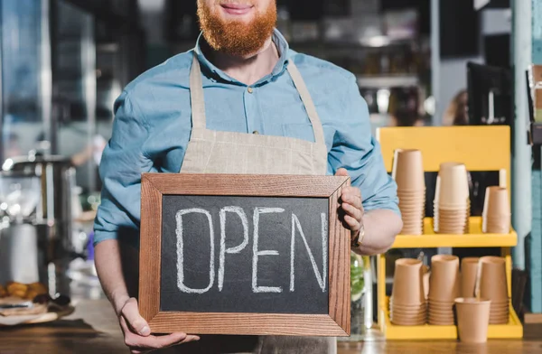 Foto recortada de un joven barista sosteniendo pizarra con letras abiertas en la cafetería - foto de stock