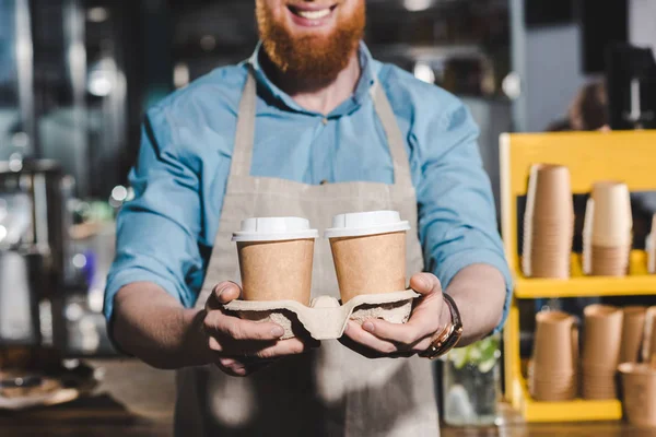 Обрезанное изображение улыбающегося мужчины-баристы с двумя одноразовыми чашками кофе — стоковое фото