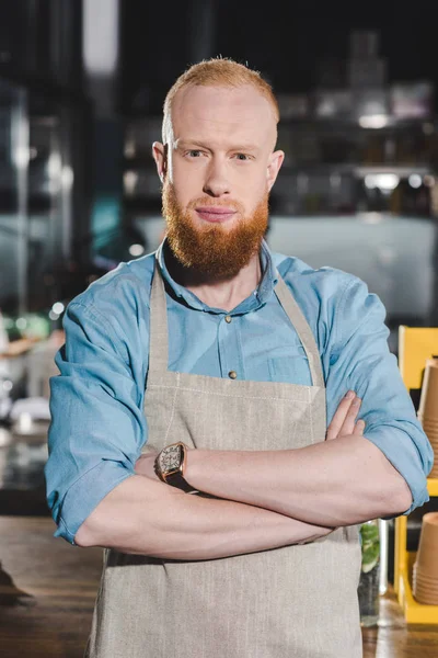 Young male barista in apron standing with crossed arms in coffee shop — Stock Photo