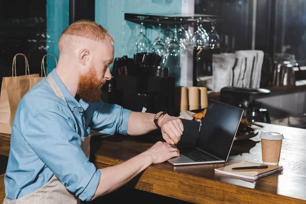 Молодой бариста смотрит на наручные часы и сидит за столом с ноутбуком в кафе — стоковое фото