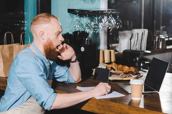 Young male barista yawning and hiding mouth by hand at table with textbook and laptop — Stock Photo