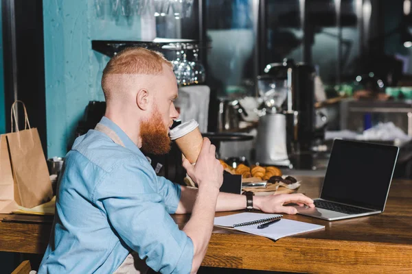 Vista laterale del giovane barista maschio che beve caffè dalla tazza di carta e utilizza il computer portatile — Foto stock