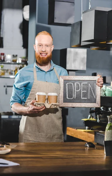 Jovem barista masculino segurando quadro com letras abertas e dois papéis xícaras de café — Fotografia de Stock