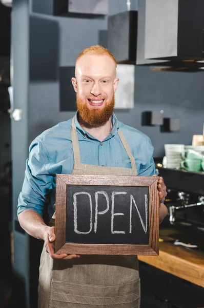 Sonriente joven barista macho sosteniendo pizarra con letras abiertas en la cafetería - foto de stock