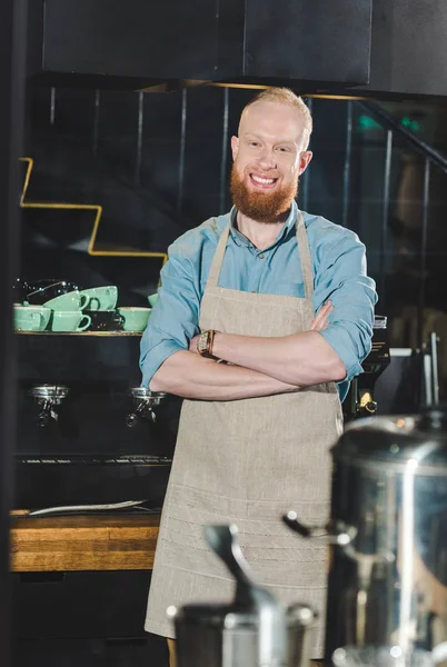 Young male barista in apron standing with crossed arms in coffee shop — Stock Photo