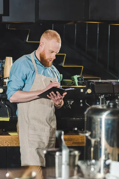 Side view of young male barista in apron writing in textbook — Stock Photo