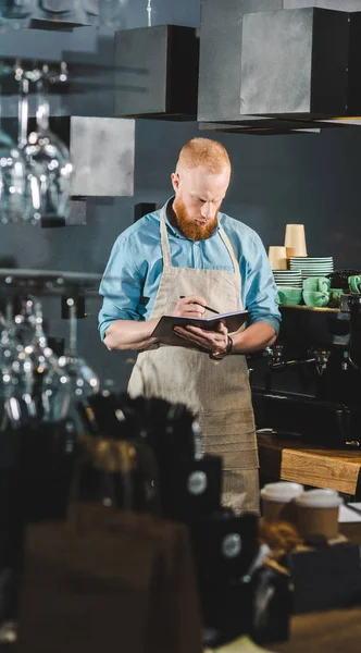 Vista frontale del giovane barista maschio in grembiule che scrive nel libro di testo — Foto stock