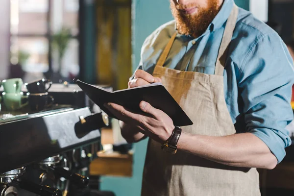 Cropped image of young male barista in apron writing in textbook — Stock Photo