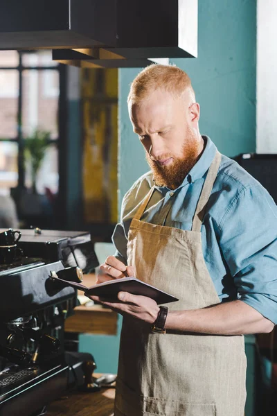 Young male barista in apron writing in textbook — Stock Photo