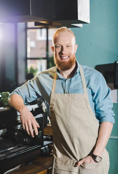 Jovem barbudo sorridente masculino barista em pé avental perto de máquina de café — Fotografia de Stock