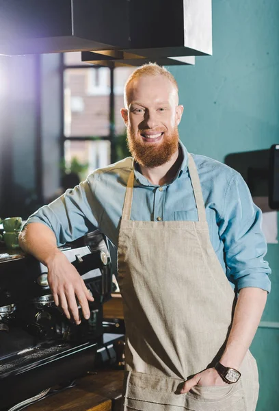 Joven barbudo sonriente barista masculino en delantal de pie cerca de la máquina de café - foto de stock