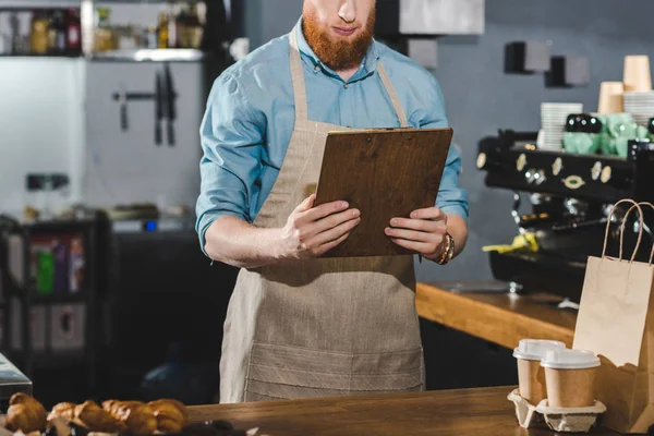 Cropped shot of serious young bearded barista holding clipboard in coffee shop — Stock Photo