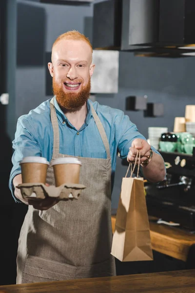 Smiling bearded barista holding paper bag and disposable coffee cups — Stock Photo