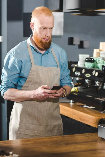 Barbudo joven barista en delantal con teléfono inteligente y mirando hacia otro lado en la cafetería - foto de stock