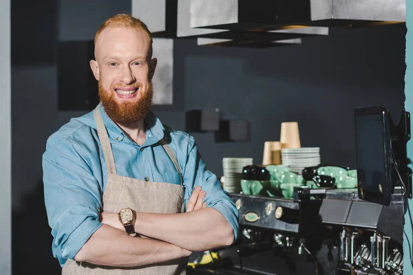 Confident bearded barista in apron standing with crossed arms and smiling at camera in coffee shop — Stock Photo