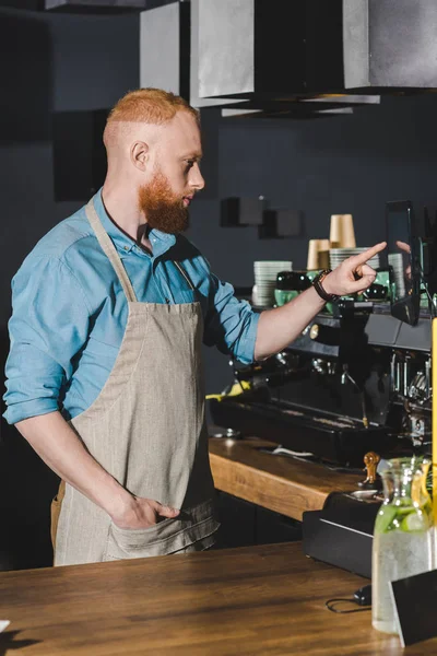 Side view of handsome young barista pointing at screen while working in coffee shop — Stock Photo