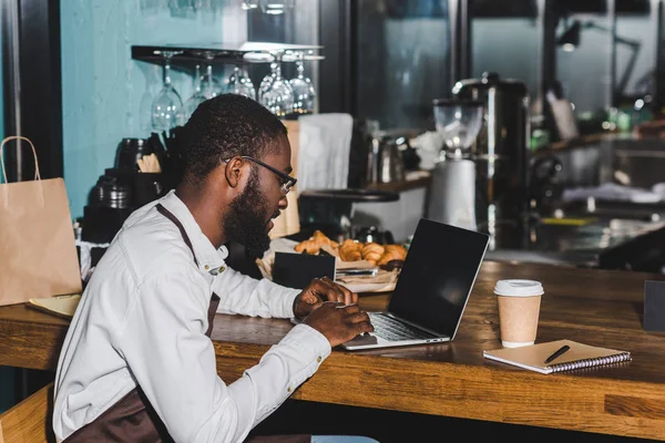 Souriant jeune afro-américaine barista dans les lunettes à l'aide d'un ordinateur portable dans un café — Photo de stock