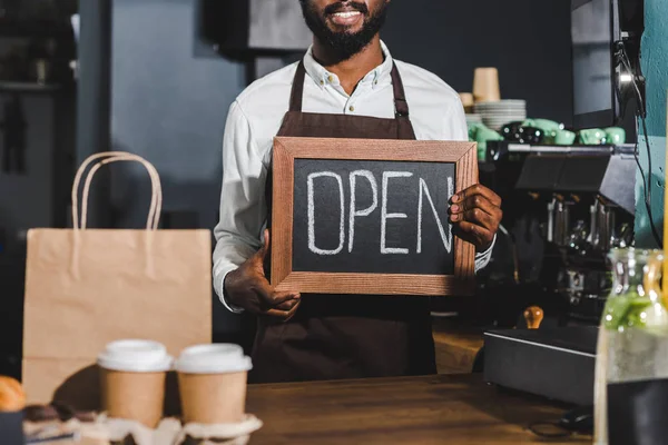Foto recortada de sonriente barista afroamericano sosteniendo cartel abierto en la cafetería - foto de stock