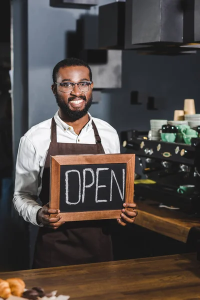 Bonito jovem africano americano barista em óculos segurando sinal aberto e sorrindo para a câmera no café — Fotografia de Stock