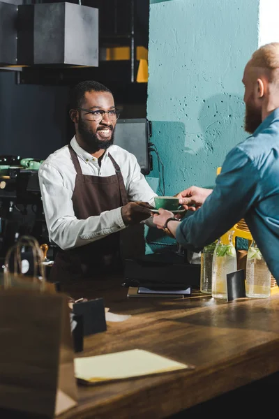 Cropped shot of cheerful african american barista giving cup of coffee to bearded young client in cafe — Stock Photo