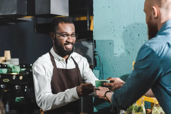 Cropped shot of smiling african american barista giving cup of coffee to bearded client in coffee shop — Stock Photo
